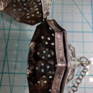 May include: A silver tea infuser with a hinged, perforated, hexagonal-shaped body. The metal surface features intricate cut-out patterns and the stamped text STERLING. A small metal chain is attached to the side of the infuser for handling.
