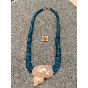 May include: A necklace featuring a long strand of small, flat, circular turquoise blue beads and a large, iridescent white conch shell pendant at the centre. A small square tag with a black cat illustration and the text BLACK CAT is included.