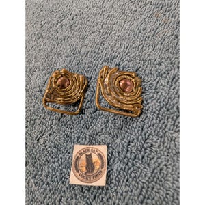 May include: A pair of gold-toned metal earrings with a swirling, textured design and a central round copper-colored accent. Below the earrings is a small square white tag featuring a black cat illustration and the text Black Cat Lucky Finds.