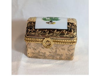 May include: A small rectangular trinket box with a hinged lid. The box features a gold-toned metal body with an embossed floral pattern. The lid has a white ceramic insert decorated with a small green tree illustration and a gold metal latch.
