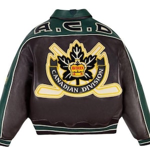 Puede incluir: Una chaqueta universitaria de cuero marrón oscuro y verde con un gran parche bordado en la espalda que muestra una hoja de arce, palos de hockey cruzados y el texto AVIREX CANADIAN DIVISION. La parte superior de la espalda lleva las letras A.C.D.