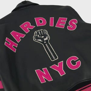 May include: A black jacket featuring a large embroidered graphic on the back. The design includes the text HARDIES in bright pink letters arched above a black and white fist icon, with the text NYC in pink letters positioned below the fist.