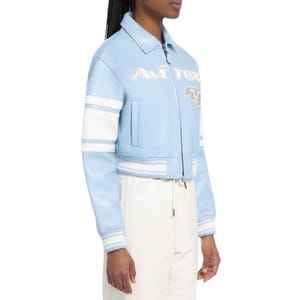 May include: A light blue leather jacket featuring a classic collar, a front zipper closure, and white stripes on the sleeves. The chest displays the embroidered text Avirex in white, accompanied by a large white and grey numerical graphic detail.