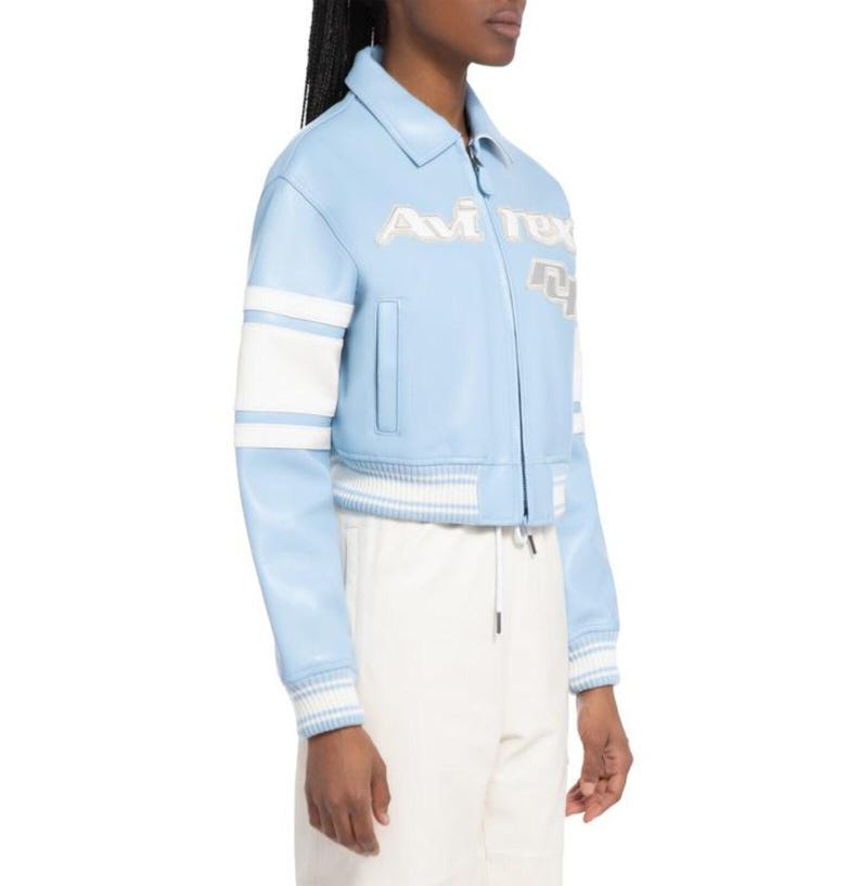 May include: A light blue leather jacket featuring a classic collar, a front zipper closure, and white stripes on the sleeves. The chest displays the embroidered text Avirex in white, accompanied by a large white and grey numerical graphic detail.