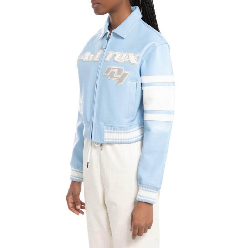 May include: A light blue varsity-style jacket featuring a white collar, white striped ribbed cuffs and hem, and white sleeve panels. The chest displays the embroidered text "Avirex" and the letters "NY" in a layered white and grey font design.