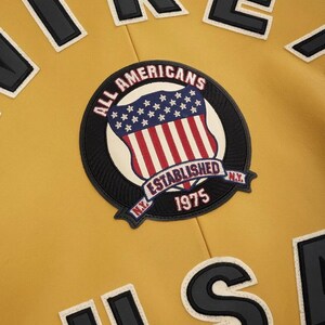 以下が含まれることがあります： マスタードイエローのジャケット。ALL AMERICANS ESTABLISHED 1975 N.Y.という文字と星条旗の盾のグラフィックが刺繍された円形のパッチが付いています。上下の端には黒と白のブロック体の文字が見えます。