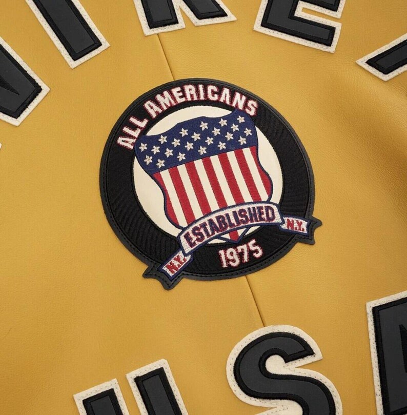 以下が含まれることがあります： マスタードイエローのジャケット。ALL AMERICANS ESTABLISHED 1975 N.Y.という文字と星条旗の盾のグラフィックが刺繍された円形のパッチが付いています。上下の端には黒と白のブロック体の文字が見えます。