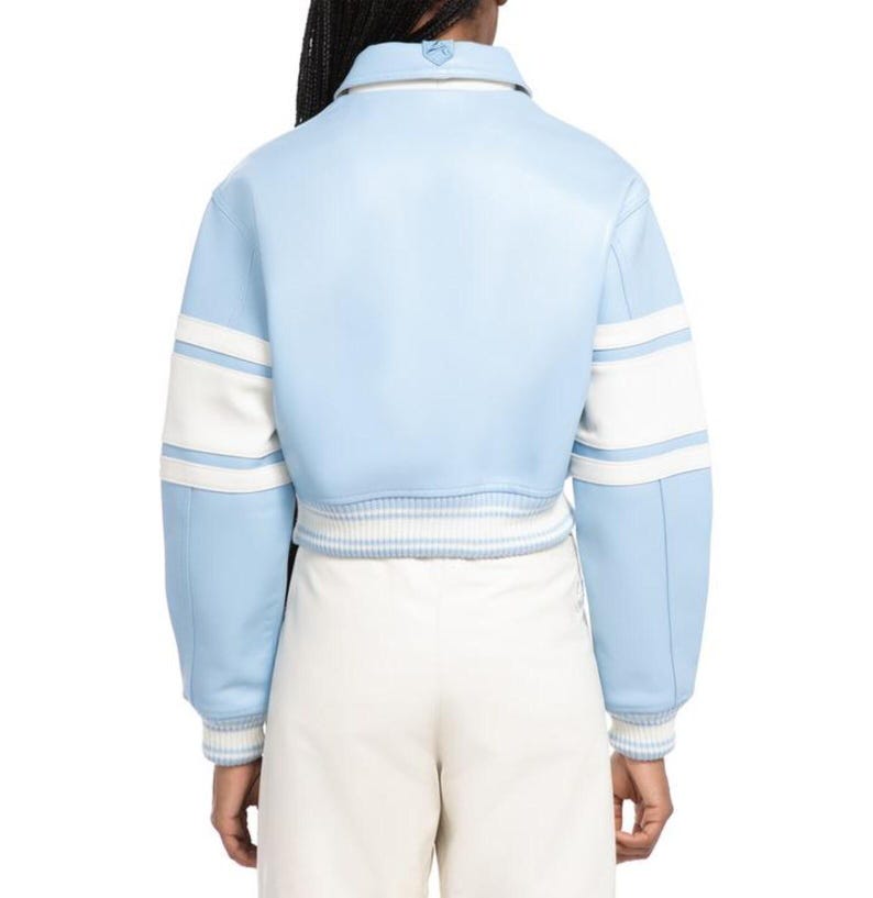 May include: A light blue varsity jacket featuring a pointed collar, long sleeves with white horizontal stripes, and a ribbed hem and cuffs with white and blue stripes. The jacket is worn over plain white trousers, showcasing a clean, structured design.