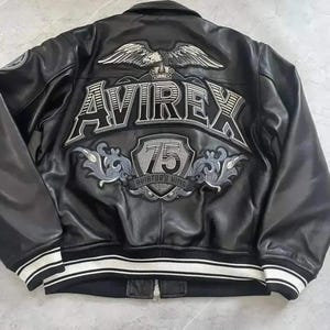 Puede incluir: Una chaqueta de cuero negra con un gran dise&ntilde;o bordado en la espalda que incluye un &aacute;guila, el texto AVIREX, un escudo con el n&uacute;mero 75 y las palabras AVIATOR'S KING. La chaqueta tiene cuello, mangas largas y un dobladillo acanalado a rayas.