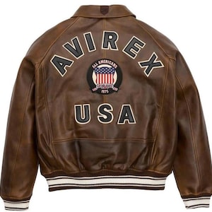 K&ouml;nnte beinhalten: Eine braune Leder-Bomberjacke mit einem gro&szlig;en, bogenf&ouml;rmigen AVIREX-Logo in schwarzer und wei&szlig;er Schrift auf der R&uuml;ckseite. Unter dem Logo befindet sich ein runder Aufn&auml;her mit US-Flagge und dem Text ALL AMERICANS ESTABLISHED 1975 USA.