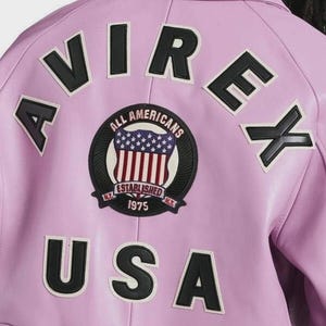 May include: A light purple jacket featuring the black text AVIREX arched across the top and USA in black letters at the bottom. A circular patch in the center displays a shield with a red, white, and blue flag design and the text ALL AMERICANS ESTABLISHED 1975 N.Y.