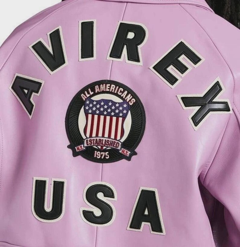 May include: A light purple jacket featuring the black text AVIREX arched across the top and USA in black letters at the bottom. A circular patch in the center displays a shield with a red, white, and blue flag design and the text ALL AMERICANS ESTABLISHED 1975 N.Y.