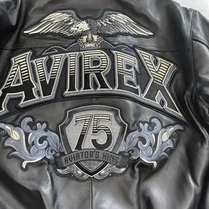 Puede incluir: Una chaqueta de cuero negra con un gran parche bordado en la espalda que muestra un &aacute;guila, una corona y el texto AVIREX. Debajo, un escudo muestra el n&uacute;mero 75 y el texto AVIATOR'S KING, rodeado de intrincados adornos en gris y negro.