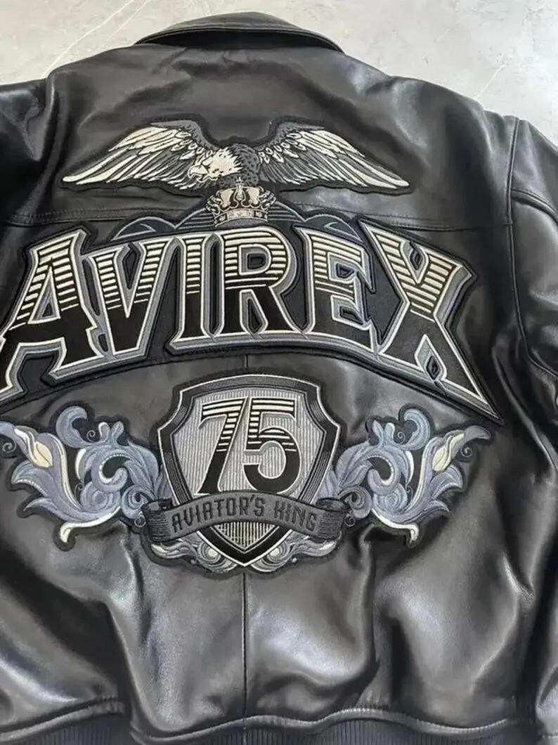 Puede incluir: Una chaqueta de cuero negra con un gran parche bordado en la espalda que muestra un &aacute;guila, una corona y el texto AVIREX. Debajo, un escudo muestra el n&uacute;mero 75 y el texto AVIATOR'S KING, rodeado de intrincados adornos en gris y negro.