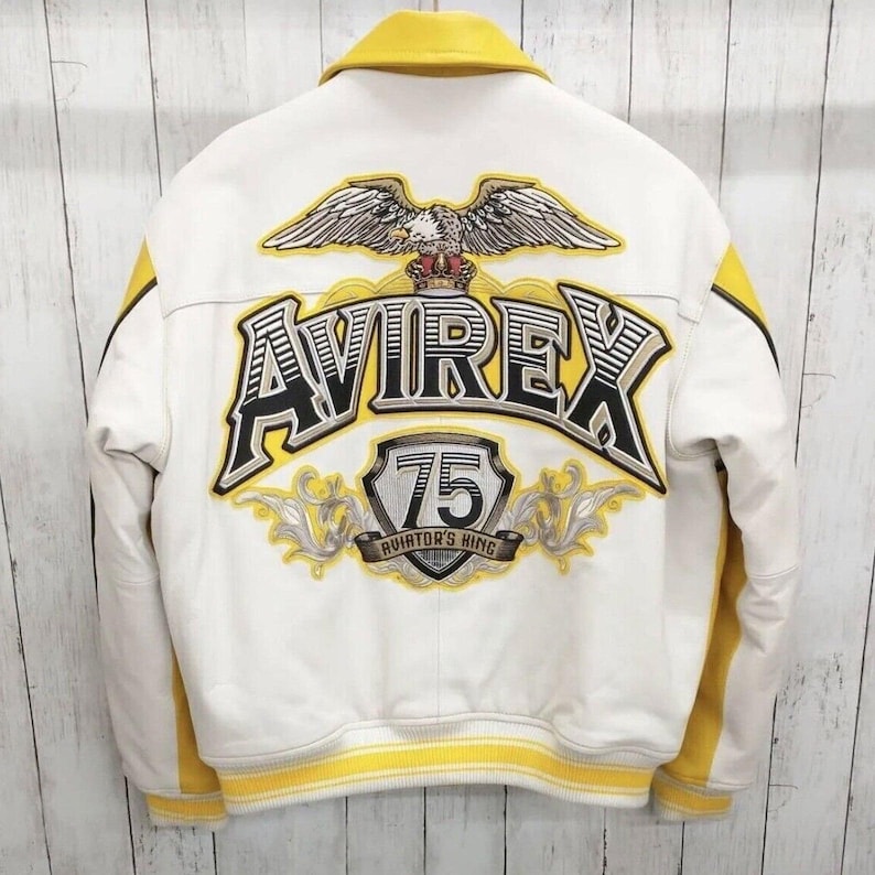Pode incluir: Uma jaqueta varsity branca e amarela com um grande gr&aacute;fico de &aacute;guia bordado nas costas. O texto AVIREX &eacute; exibido de forma proeminente acima de um emblema de escudo contendo o n&uacute;mero 75 e as palavras AVIATOR'S KING em preto e amarelo.