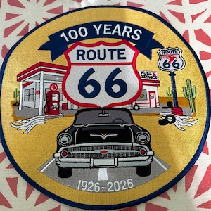 May include: A circular embroidered patch featuring a gold background with a black car driving on a road. It displays a Route 66 shield, a gas station, a Route 66 sign, and cacti. Text includes 100 YEARS, ROUTE 66, FUEL & SERVICE, and 1926-2026.