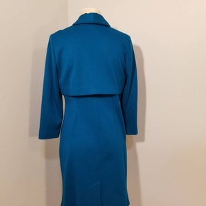 May include: A teal blue dress with a matching jacket. The dress has a fitted silhouette and a long sleeve. The jacket has a collar and a long sleeve.