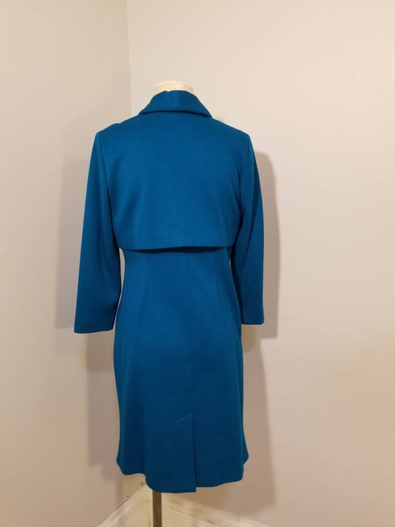 May include: A teal blue dress with a matching jacket. The dress has a fitted silhouette and a long sleeve. The jacket has a collar and a long sleeve.