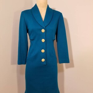 May include: A teal blue dress with a long sleeve and a collar. The dress has a row of gold buttons down the front.