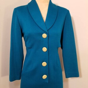 May include: A teal blue dress with a long sleeve and a collar. The dress has a row of gold buttons down the front.