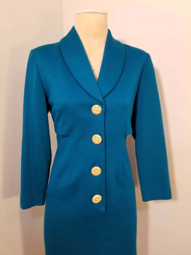 May include: A teal blue dress with a long sleeve and a collar. The dress has a row of gold buttons down the front.