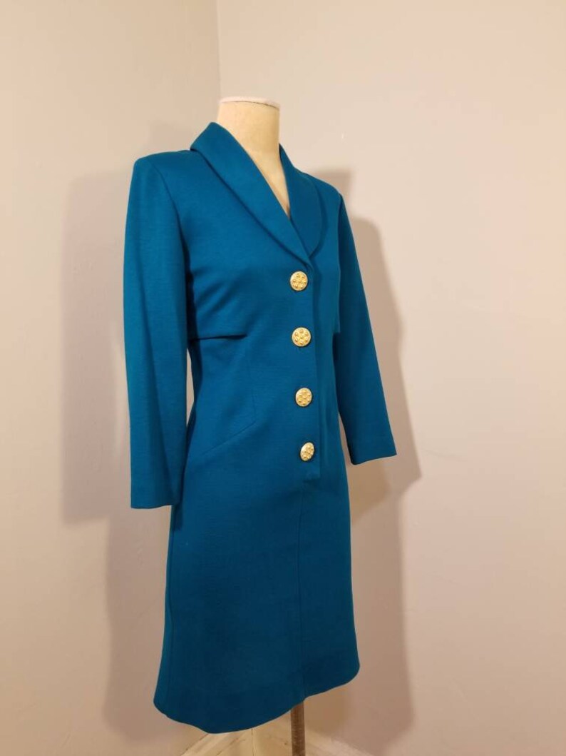 May include: Teal blue dress with a collar and gold buttons. The dress has a fitted silhouette and a knee-length hem.