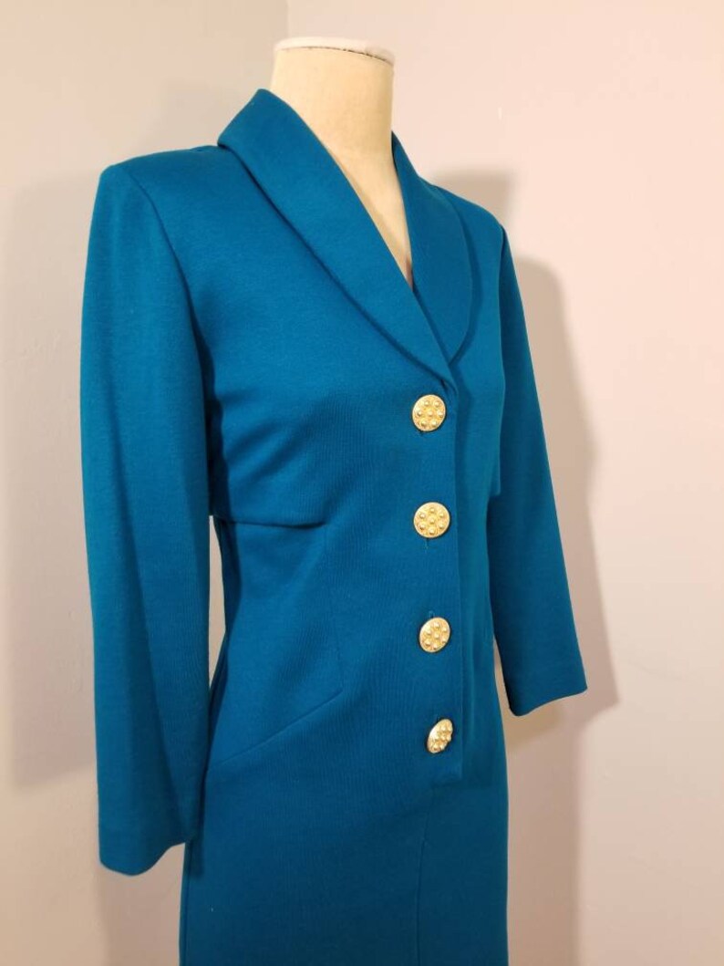 May include: A teal blue blazer with a collar and five gold buttons. The blazer is on a white mannequin.
