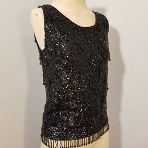 May include: A sleeveless black sequined top with a fringe trim. The top is made of small black sequins and has a scoop neckline.