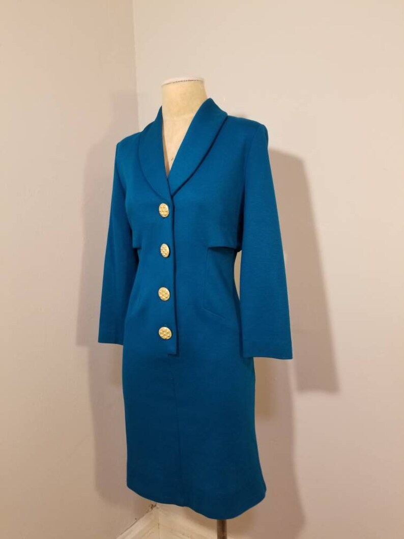 May include: A teal blue dress with a long sleeve and a collar. The dress has gold buttons down the front.