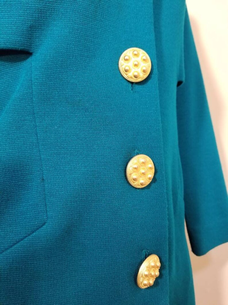 May include: Teal blue blazer with three gold tone buttons. The buttons have a raised, circular design.