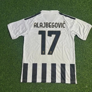 Könnte beinhalten: Ein weißes kurzärmeliges Sporttrikot mit dem Namen ALAJBEGOVIĆ und der Nummer 17 in dunkelblauer Schrift auf der Rückseite. Der untere Teil des Trikots zeigt vertikale schwarze und weiße Streifen, und die Nummer 17 enthält ein kleines Logo.