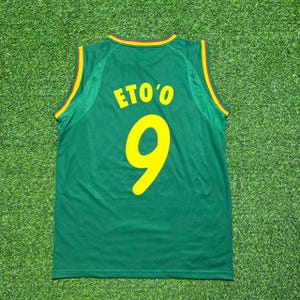 May include: A green sleeveless sports jersey featuring the name ETO'O and the number 9 printed in yellow on the back. The collar and arm openings are trimmed with thin red, yellow, and green stripes, creating a distinct athletic appearance.