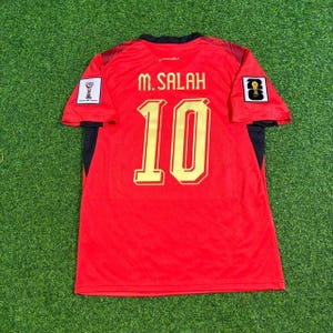 以下が含まれることがあります： 背面にゴールドの文字で「M. SALAH」と「10」がプリントされた赤いスポーツジャージ。袖には黒いアクセントとワッペンがあり、右袖にはFIFAワールドカップのエンブレム、左袖には円形のロゴがあしらわれています。