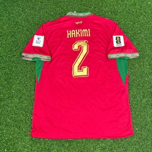 May include: A red soccer jersey featuring the name HAKIMI and the number 2 in gold lettering on the back. The sleeves have green accents with patterned trim, and the shoulders display white patches with text and logos, including 2026.
