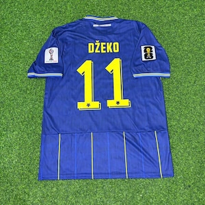 May include: A blue soccer jersey featuring the name DŽEKO and the number 11 in yellow on the back. The shirt includes a small Bosnia and Herzegovina text at the collar, a circular logo on the right sleeve, and a square 26 logo on the left sleeve.