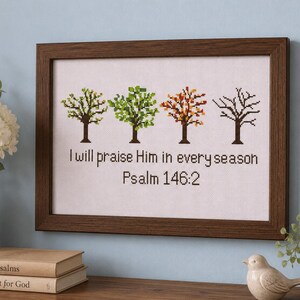 Könnte beinhalten: Ein gerahmtes Kreuzstichbild mit vier Bäumen, die verschiedene Jahreszeiten darstellen, sowie dem Text "I will praise Him in every season" und "Psalm 146:2" in schwarzem Garn auf weißem Stoff, eingefasst in einen dunkelbraunen Rahmen.