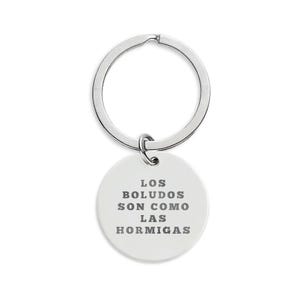 Puede incluir: Un llavero plateado con un colgante blanco y redondo. El colgante tiene el texto "LOS BOLUDOS SON COMO LAS HORMIGAS" en letras grises. El llavero está sobre un fondo blanco.
