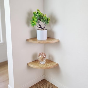 May include: Two light-coloured wooden corner shelves mounted on a white wall. The top shelf holds a small green artificial plant in a white pot. The bottom shelf holds a clear glass decorative container with a metallic finish and dried contents.