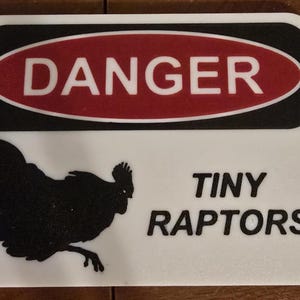 May include: A rectangular white sign with rounded corners featuring a red oval at the top containing the word DANGER in white capital letters. Below this, a black silhouette of a chicken is on the left and the text TINY RAPTORS in black.