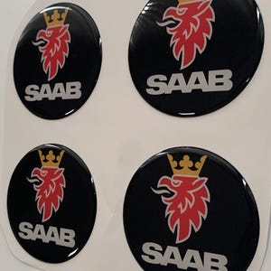 May include: Four circular domed stickers featuring the Saab logo. Each sticker has a black background with a red griffin head wearing a gold crown above the white text SAAB. The stickers are arranged in a two-by-two grid on a white backing sheet.