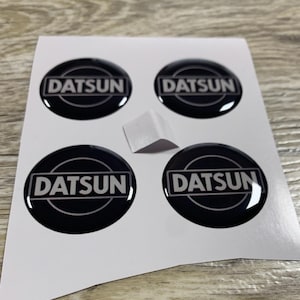 Può includere: Un set di quattro adesivi circolari neri bombati disposti su un foglio bianco. Ogni adesivo presenta un bordo circolare bianco e una barra rettangolare orizzontale centrale contenente il testo bianco DATSUN in un carattere sans-serif.