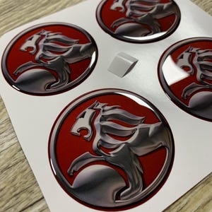 Peut inclure: Une feuille de trois autocollants circulaires présentant un emblème de lion en argent métallique sur un fond rouge uni. Chaque autocollant a une finition bombée brillante, représentant une figure de lion stylisée de profil dans un cercle.