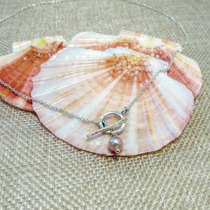 May include: A silver necklace with a toggle clasp and a single pink pearl pendant. The necklace is displayed on a bed of seashells.