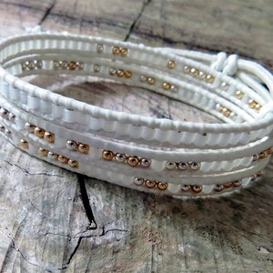May include: A white leather wrap bracelet with gold and silver beads. The bracelet is made of multiple strands of leather that are woven together.