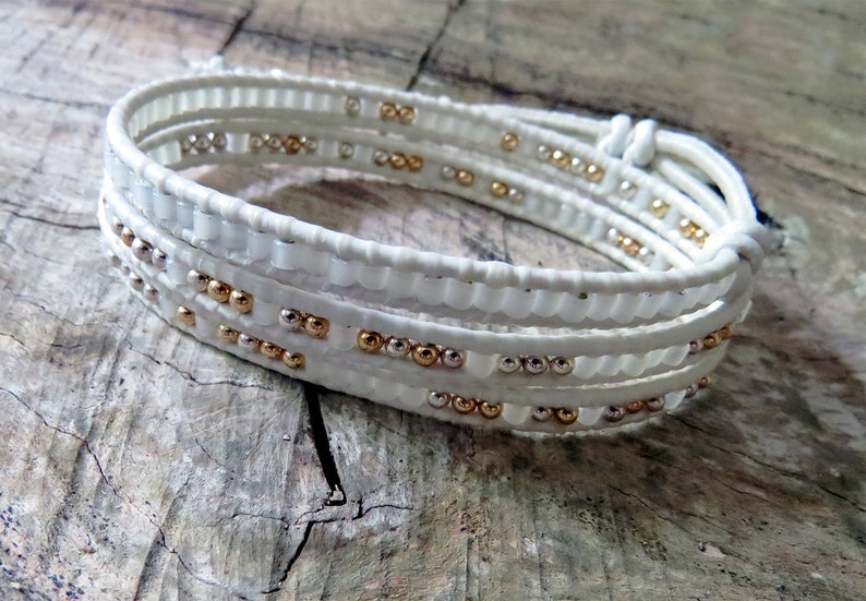 May include: A white leather wrap bracelet with gold and silver beads. The bracelet is made of multiple strands of leather that are woven together.