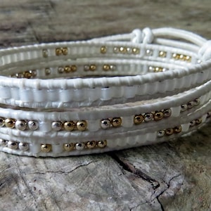 Puede incluir: Pulsera de cuero blanco con cuentas doradas y plateadas. La pulsera está hecha de múltiples hebras de cuero que se envuelven alrededor de la muñeca.