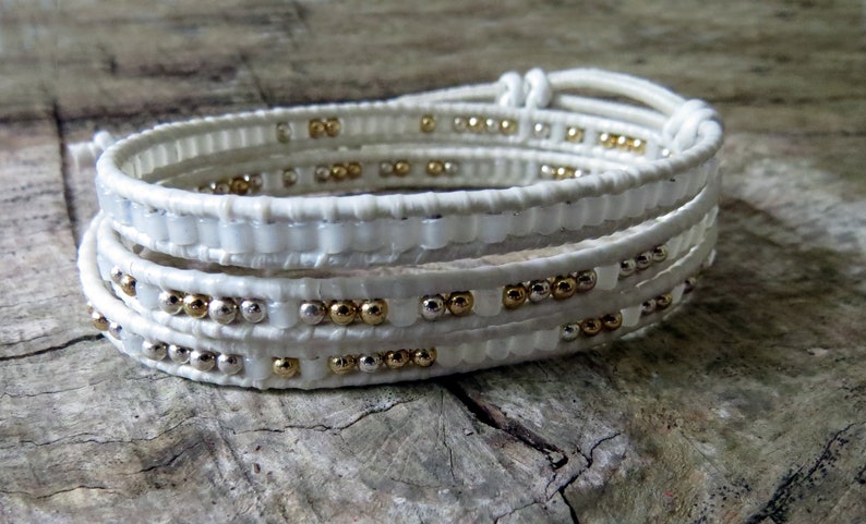 May include: White leather wrap bracelet with gold and silver beads. The bracelet is made of multiple strands of leather that are wrapped around the wrist.