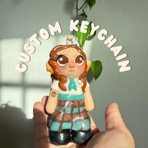 Puede incluir: Un llavero personalizado con una figura de arcilla pintada a mano con cabello castaño, ojos grandes con reflejos de estrellas y pecas. La figura lleva una camisa blanca con corbata turquesa, una etiqueta con el nombre TAI y una falda de cuadros.