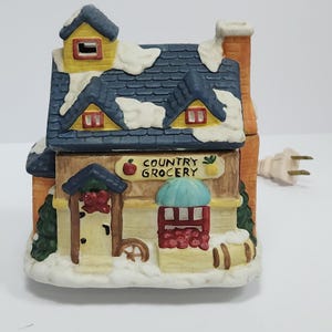 May include: A ceramic miniature building shaped like a Country Grocery store with a blue shingled roof covered in white snow. The structure features a yellow dormer, a brick chimney, a front door with a red wreath, and a window displaying red produce.