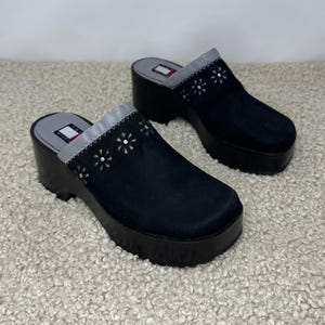 May include: A pair of black platform clogs featuring a thick black sole and a suede upper. The top of the shoe is decorated with a row of white flower-shaped cutouts with silver studs in the center. The insole is grey with a Tommy Hilfiger logo.